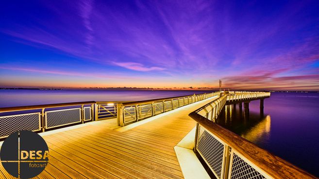 Ein beleuchteter Steg erstreckt sich über ruhiges Wasser unter einem farbenfrohen Abendhimmel.