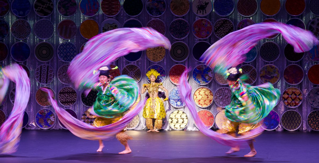 Two dancers in colourful costumes whirl bright ribbons, with one person in gold costume standing behind them in front of circular ornaments.