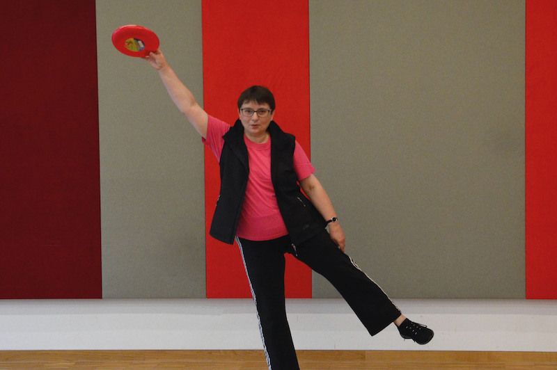 Eine Frau in sportlicher Kleidung hält eine rote Frisbee und balanciert auf einem Bein vor einer bunten Wand.