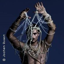 Ein Tänzer, der mit Perlenketten bedeckt ist, hebt die Arme vor einem dunklen Hintergrund.