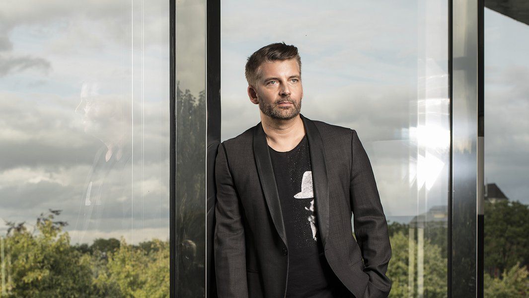 Ein Mann in schwarzem Anzug steht vor einer Glaswand mit Blick auf eine bewölkte Landschaft.