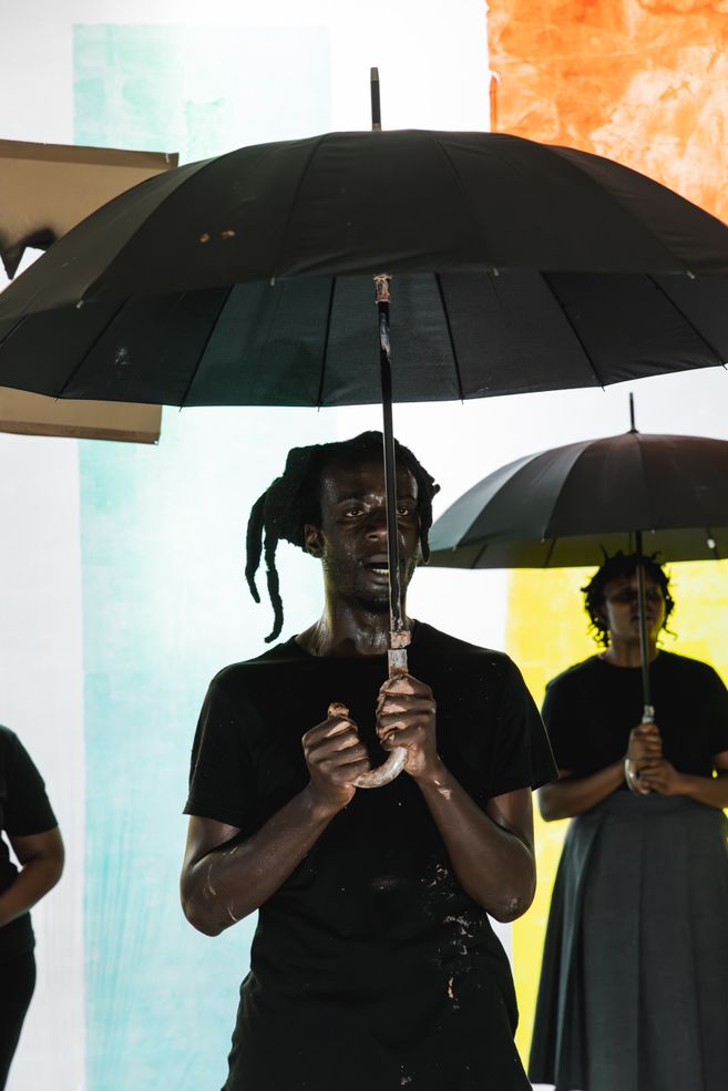 Excerpt from the production DAMBUDZO by nora chipaumire. A person stands holding an umbrella in front of a colorful wall. Behind them, two other people are visible in the background.