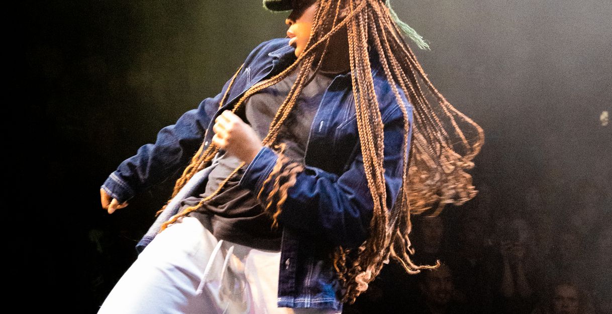A person wearing a knitted hat, long braids, and loose clothing stands on a stage and moves their hips in one direction with bent knees. They pull themselves up in the same direction with the opposite arm.