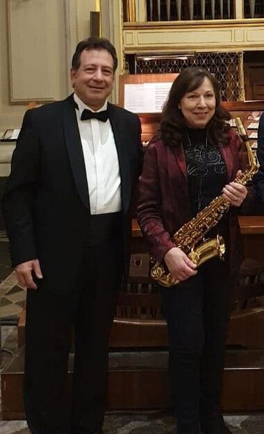 Ein Mann im Smoking und eine Frau mit Saxophon stehen vor einer Orgel in einem eleganten Raum.