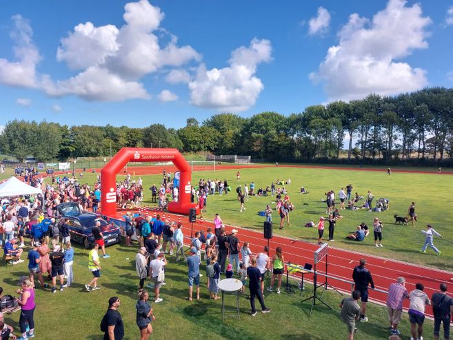 Menschenmenge bei einem Marathon-Event auf einem Sportplatz mit rotem Startbogen der Sparkasse Holstein.