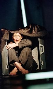 Eine Person kauert in einem offenen Koffer, trägt einen Anzug und hat einen besorgten Gesichtsausdruck.