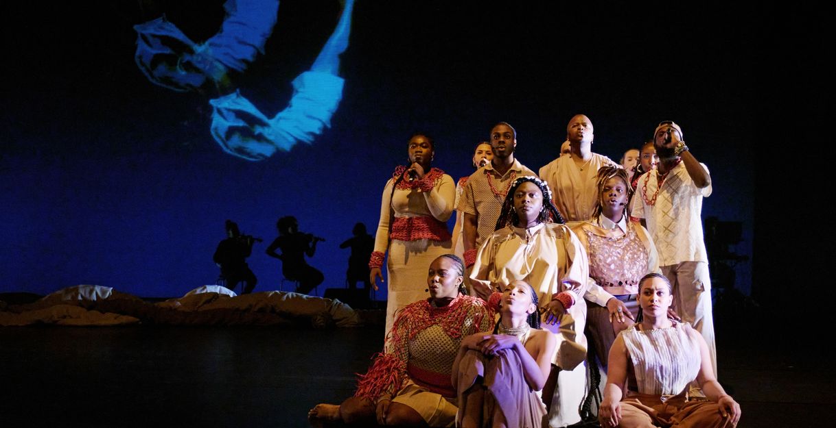 A group of performers in beige and red costumes sit and stand in formation on stage. In the background, silhouettes of musicians and a projection of a floating body are visible.
