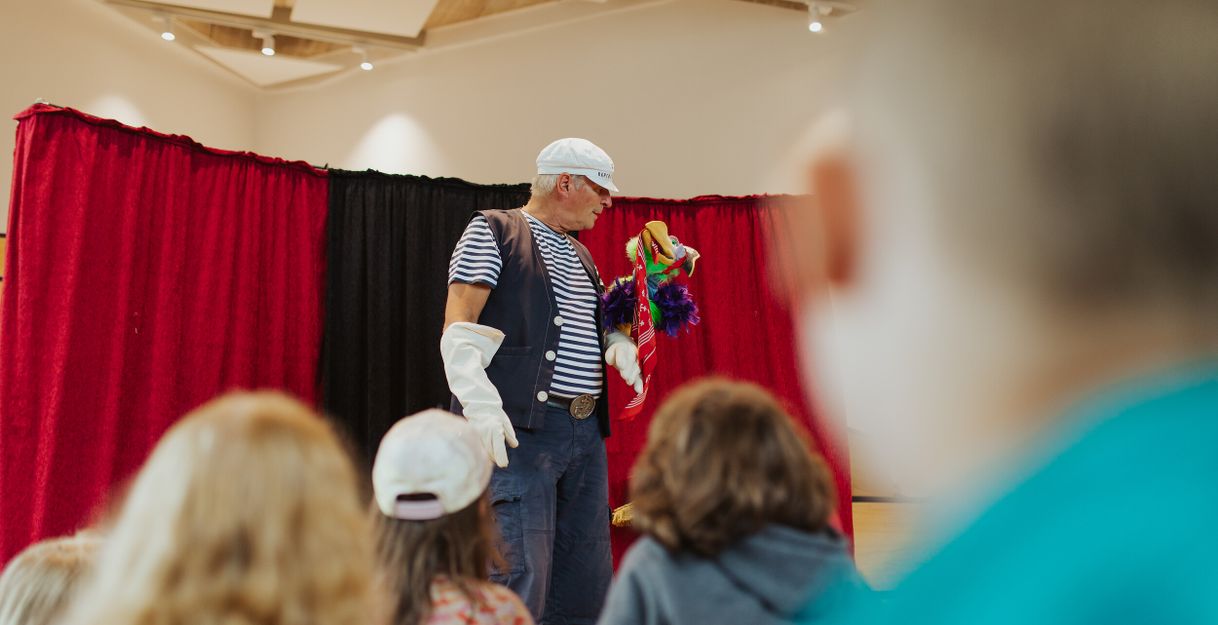Ein Mann in Matrosenkleidung führt eine Handpuppe vor einem roten Vorhang vor Kindern vor.