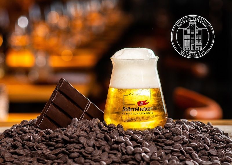 Ein Glas Bier der Marke Störtebeker steht auf einem Haufen Schokoladenstückchen, daneben eine Tafel Schokolade. Im Hintergrund das Logo von Evers & Tochter Manufaktur.