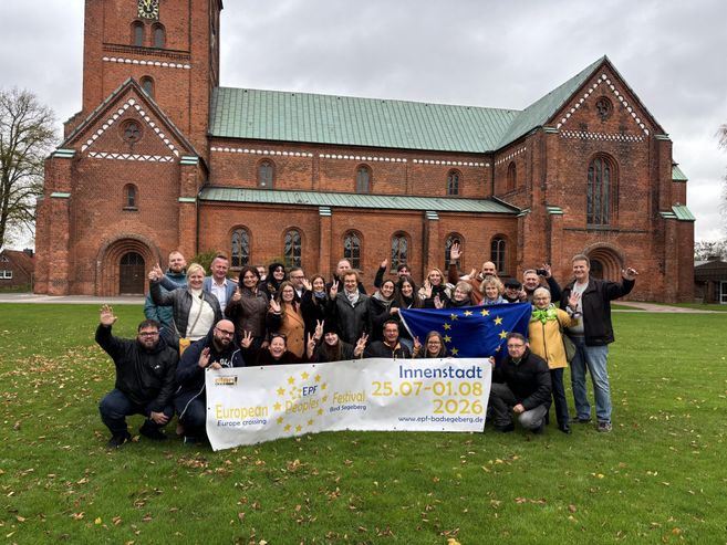 Eine Gruppe von Menschen steht vor einer Kirche und hält ein Banner hoch. Der Banner zeigt Informationen zu einer Veranstaltung in der Innenstadt.