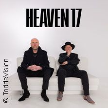 Zwei Männer sitzen auf einem weißen Sofa vor einem weißen Hintergrund mit dem Schriftzug 'Heaven 17'.