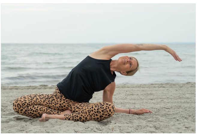Eine Person macht Yoga am Strand, sitzt im Schneidersitz und streckt den Oberkörper zur Seite. Im Hintergrund ist das Meer zu sehen.