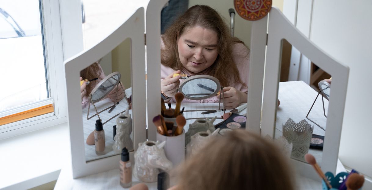 Eine hellhäutige, junge Frau sitzt an einem weißen Schminktisch, auf dem ein großer, dreiteiliger Spiegel montiert ist. Der Tisch steht vor einem Fenster.  Auf dem Schminktisch stehen verschiedene kosmetische Produkte, die für ein Make-up benötigt werden – z.B. Schwämmchen, Pinsel und Puder. Die verschiedenen Pinsel stecken in dekorativen kleinen Bechern. Die Frau hält einen Mascara-Pinsel und einen Kajalstift in der Hand. Sie ist offenbar dabei, ihre Augen zu tuschen.  Das Foto ist vom Rücken der Frau aus aufgenommen. Man erkennt ihr konzentriertes Gesicht von vorne im großen mittleren Spiegel des Schminktischs. Sichtbar ist in dem großen Spiegel auch, dass vor ihr auf dem Tisch zwischen den Schminksachen auch noch ein kleiner runder Aufstellspiegel steht, auf den sie gerade schaut. Hinter ihrem Rücken erkennt man den Griff ihres Rollstuhls.  Die Frau ist etwa 25 Jahre alt, sie hat dunkelblonde Haare, die offen auf ihren Schultern liegen. Sie hat ein rosa Kapuzen-Sweatshirt an.