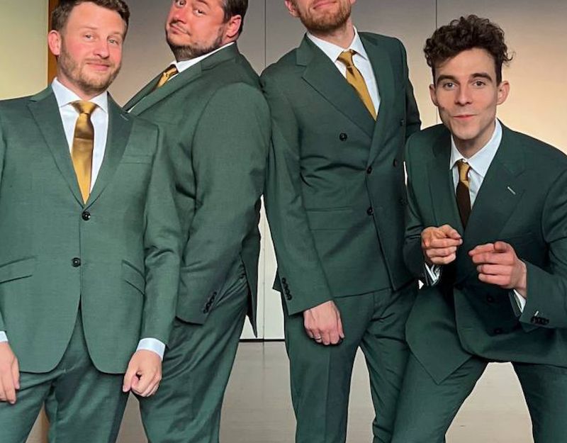 Vier Männer in grünen Anzügen mit goldenen Krawatten posieren lächelnd.