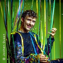 Ein Mann in einem bunten Outfit sitzt in einem Raum voller bunter Bänder und Konfetti.