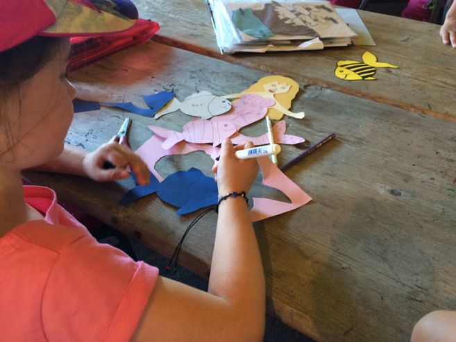 Ein Kind bastelt aus buntem Papier eine Meerjungfrau an einem Holztisch.