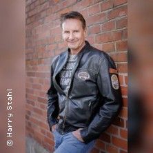 Ein Mann in Lederjacke lehnt an einer Backsteinmauer.