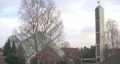 Kirche mit spitzem Dach und Glockenturm in einer bewaldeten Umgebung.