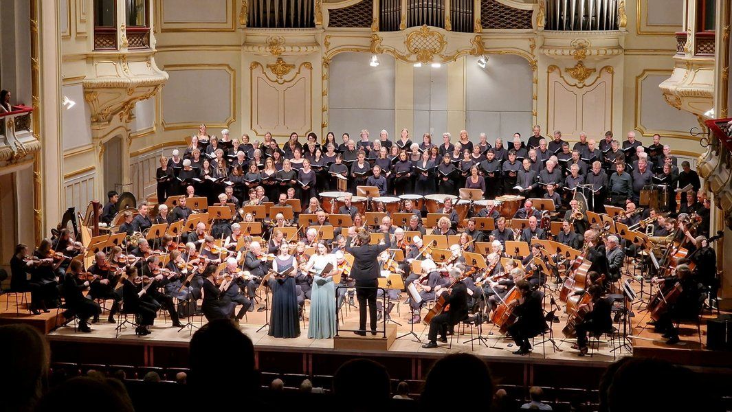 Ein Orchester und Chor führen in einem prunkvollen Konzertsaal ein klassisches Musikstück auf.