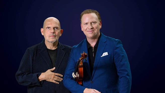 Zwei Männer in formeller Kleidung vor einem dunkelblauen Hintergrund, einer hält eine Geige.