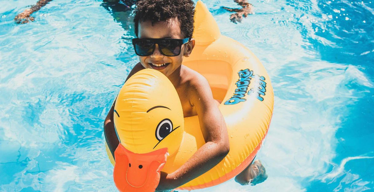 Ein lachendes Kind mit Sonnenbrille sitzt in einem gelben Schwimmring in Form einer Ente im Pool.