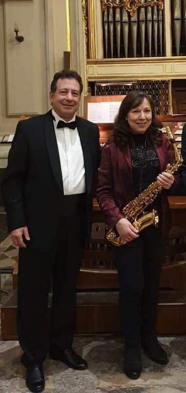 Ein Mann im Smoking und eine Frau mit Saxophon stehen vor einer Orgel in einem eleganten Raum.