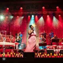 Eine Sängerin in einem rosa Kleid tritt auf einer Bühne mit Band und roter Beleuchtung auf.