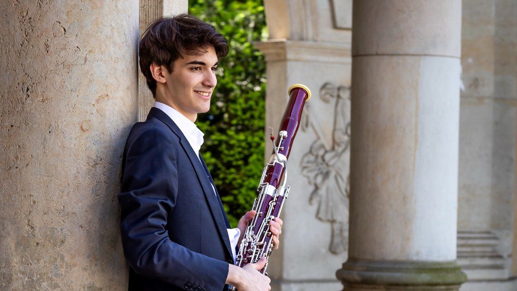 Ein junger Mann in einem Anzug lehnt an einer Säule und hält ein Fagott.