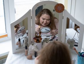 Eine hellhäutige, junge Frau sitzt an einem weißen Schminktisch, auf dem ein großer, dreiteiliger Spiegel montiert ist. Der Tisch steht vor einem Fenster.  Auf dem Schminktisch stehen verschiedene kosmetische Produkte, die für ein Make-up benötigt werden – z.B. Schwämmchen, Pinsel und Puder. Die verschiedenen Pinsel stecken in dekorativen kleinen Bechern. Die Frau hält einen Mascara-Pinsel und einen Kajalstift in der Hand. Sie ist offenbar dabei, ihre Augen zu tuschen.  Das Foto ist vom Rücken der Frau aus aufgenommen. Man erkennt ihr konzentriertes Gesicht von vorne im großen mittleren Spiegel des Schminktischs. Sichtbar ist in dem großen Spiegel auch, dass vor ihr auf dem Tisch zwischen den Schminksachen auch noch ein kleiner runder Aufstellspiegel steht, auf den sie gerade schaut. Hinter ihrem Rücken erkennt man den Griff ihres Rollstuhls.  Die Frau ist etwa 25 Jahre alt, sie hat dunkelblonde Haare, die offen auf ihren Schultern liegen. Sie hat ein rosa Kapuzen-Sweatshirt an.