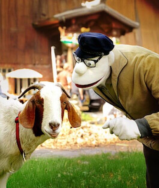 Eine Puppe in einer Jacke und Mütze beugt sich zu einer Ziege auf einer Wiese vor einem Holzgebäude.