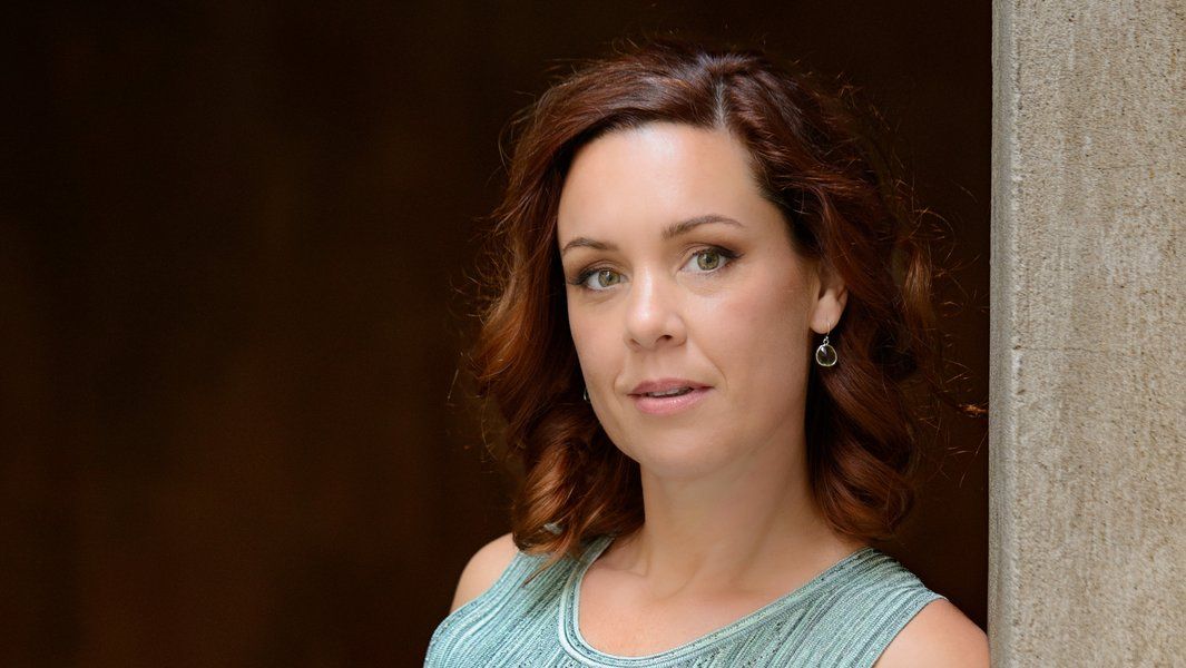 Porträt einer Frau mit braunen Haaren und einem blauen Kleid vor einem dunklen Hintergrund.