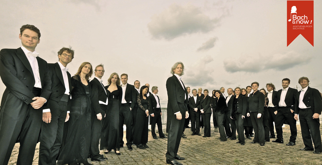 Eine Gruppe von Musikern in formeller Kleidung steht auf einem gepflasterten Platz unter einem bewölkten Himmel.