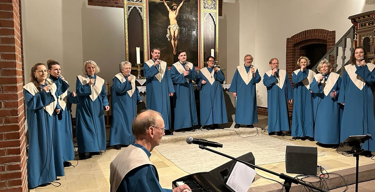 Ein Gospelchor in blauen Roben singt in einer Kirche vor einem Altarbild. Ein Mann spielt Klavier im Vordergrund.