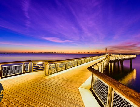 Ein beleuchteter Steg erstreckt sich über ruhiges Wasser unter einem farbenfrohen Abendhimmel.