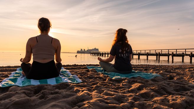 Zwei Personen meditieren am Strand bei Sonnenuntergang mit einem Schiff im Hintergrund.