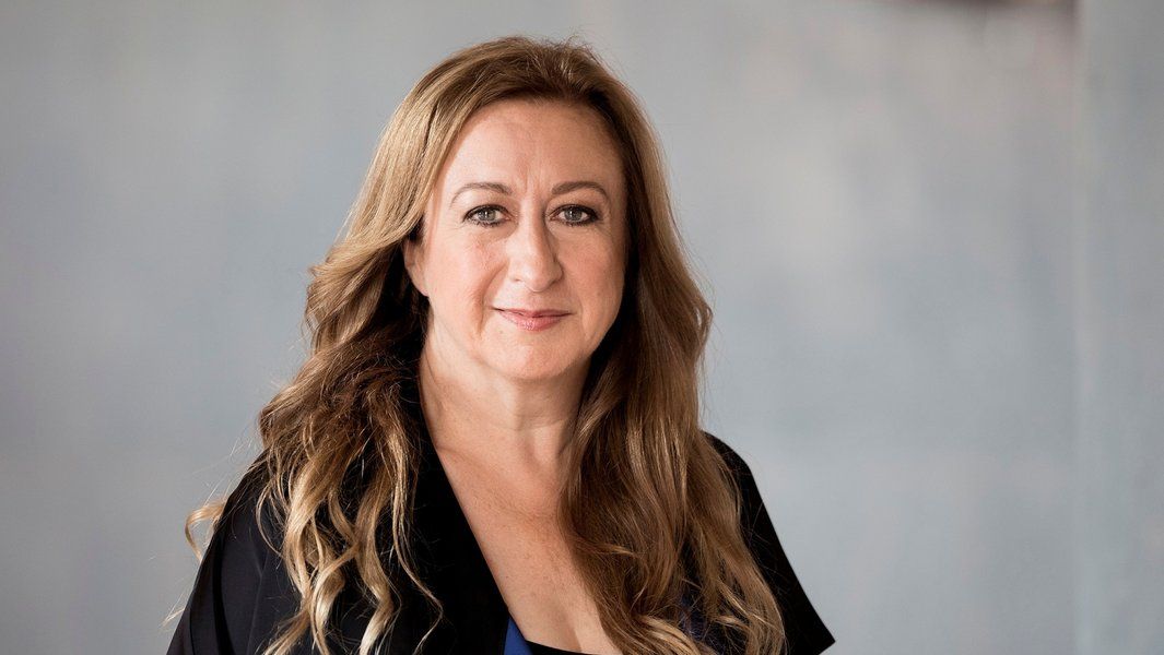 Porträt einer Frau mit langen, welligen Haaren und einem schwarzen Oberteil vor einem neutralen Hintergrund.