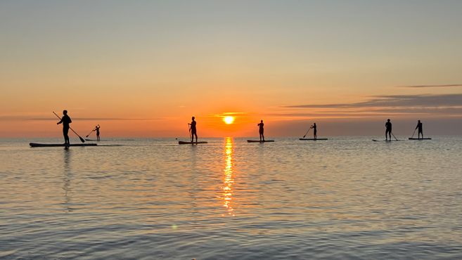 Personen beim Stand-Up-Paddling bei Sonnenaufgang auf dem Meer.