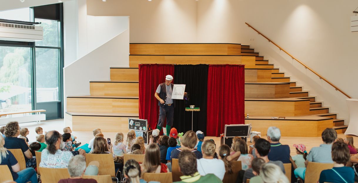 Ein Zauberer in Matrosenkleidung führt vor einem Publikum von Kindern und Erwachsenen auf einer Bühne mit roten Vorhängen eine Show auf.