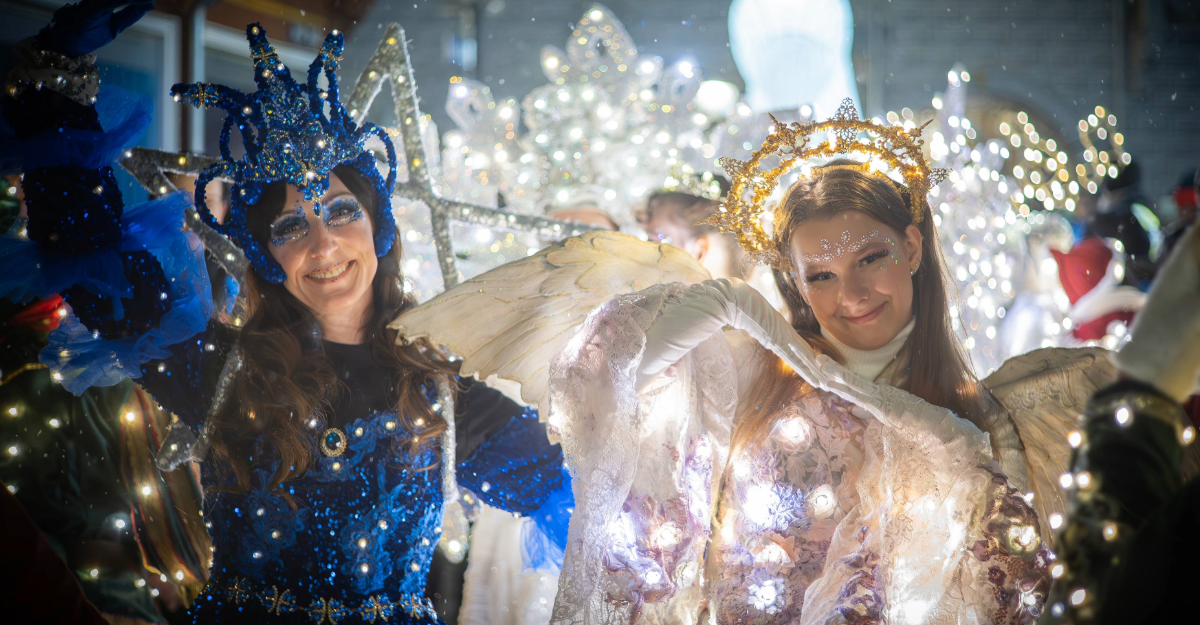 Zwei Frauen in funkelnden Kostümen mit Lichtern, eine in Blau, die andere in Weiß mit Flügeln, lächeln in die Kamera.