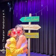 Zwei Personen mit roten Nasen umarmen sich vor einem Wegweiser auf einer Bühne.