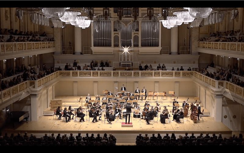 Orchester spielt in einem großen Konzertsaal mit Publikum auf Balkonen.