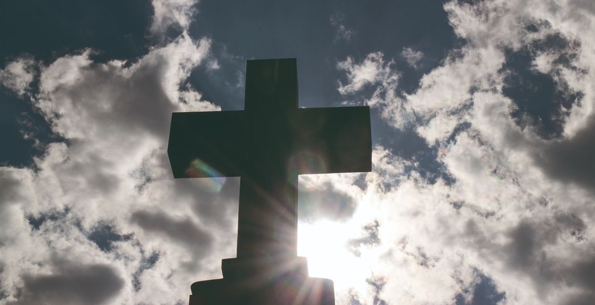 Ein Kreuz vor einem bewölkten Himmel mit Sonnenstrahlen.