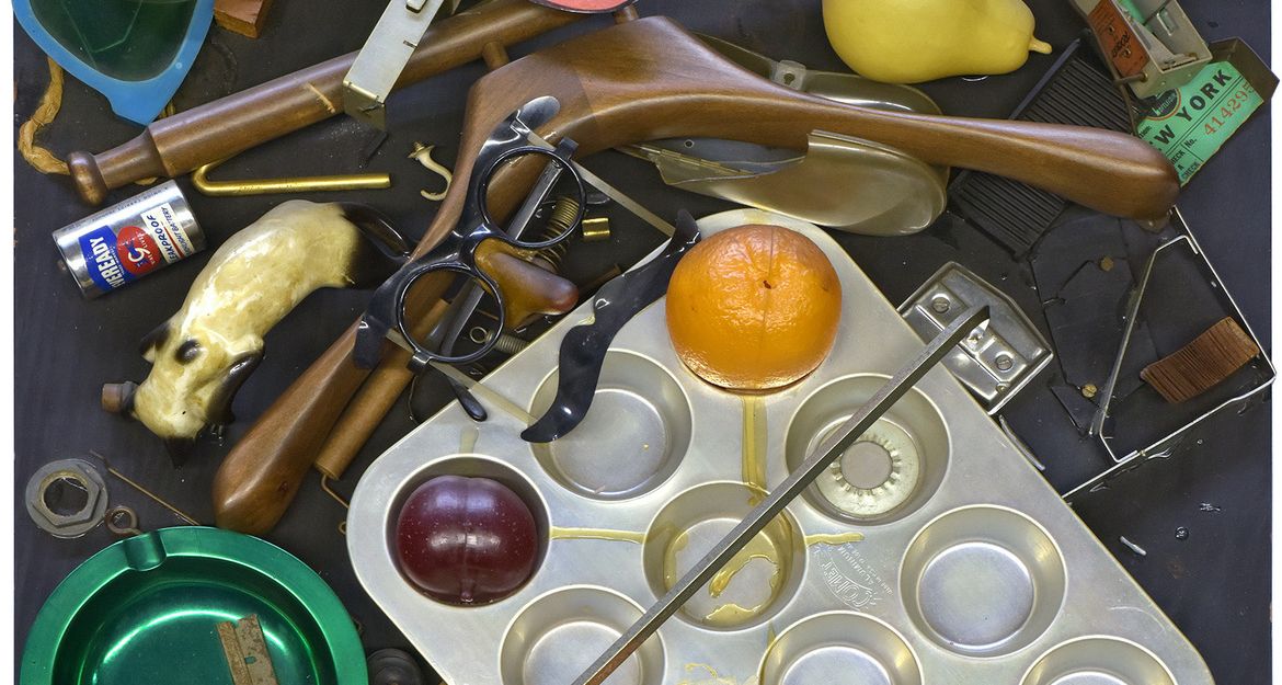 Ein Kunstwerk von Daniel Spoerri mit verschiedenen Alltagsgegenständen wie Brille, Obst, Backform und Spielzeugpistole auf einem Tisch arrangiert.