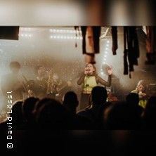 Eine Band spielt auf einer Bühne vor Publikum, beleuchtet von warmem Licht.