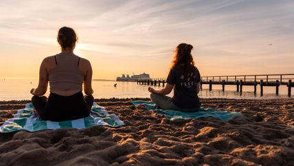 Zwei Personen meditieren am Strand bei Sonnenuntergang mit einem Schiff im Hintergrund.