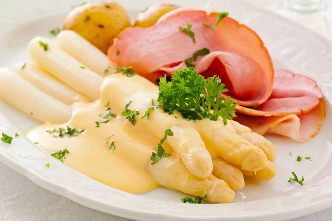 Ein Teller mit weißem Spargel, Schinken, Kartoffeln und Sauce Hollandaise, garniert mit Petersilie.