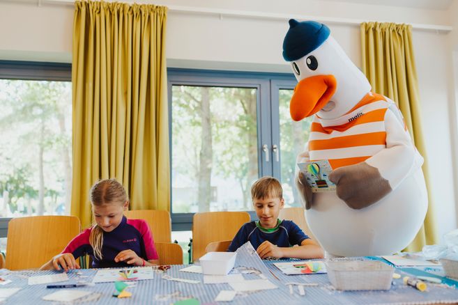 Zwei Kinder basteln an einem Tisch, während ein großes Möwenmaskottchen daneben steht.