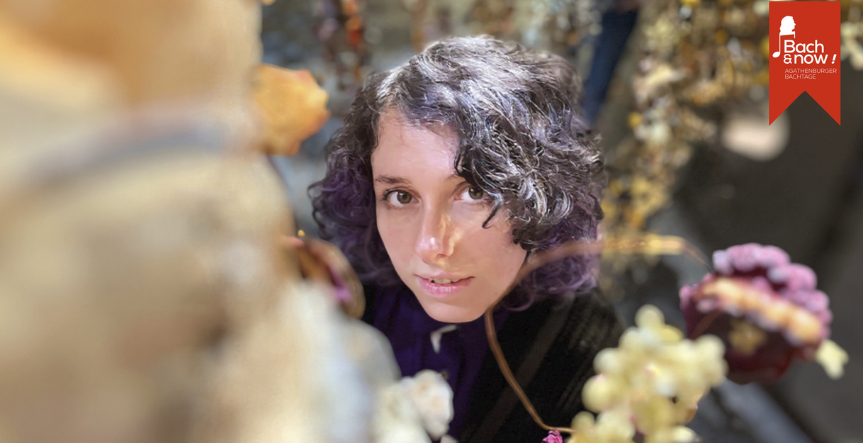 Eine Frau mit lockigem Haar schaut durch einen Blumenrahmen in die Kamera. Im Hintergrund ist ein Banner mit der Aufschrift 'Bach & now! Agathenburger Bachtage'.