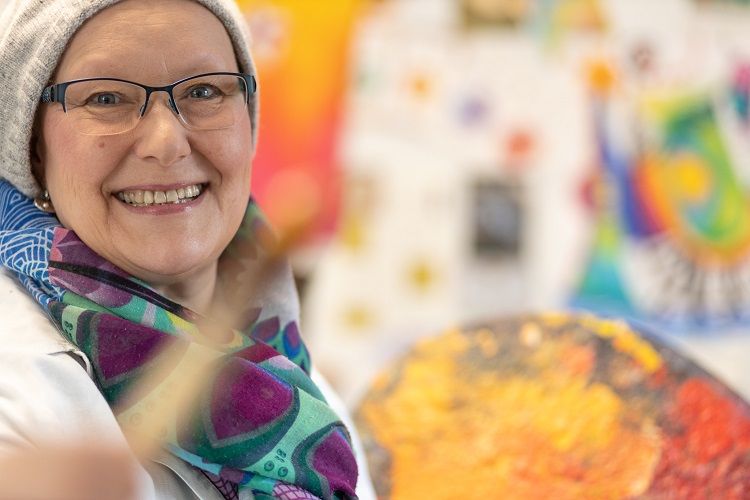 Frau mit Brille und Mütze lächelt in die Kamera, hält einen Pinsel. Hintergrund zeigt bunte Malerei.