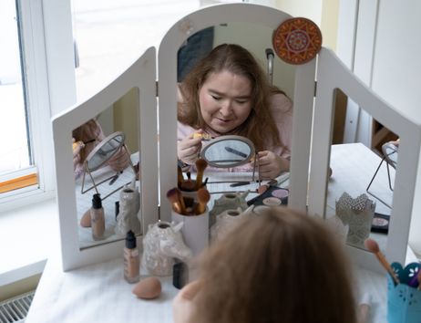 Eine hellhäutige, junge Frau sitzt an einem weißen Schminktisch, auf dem ein großer, dreiteiliger Spiegel montiert ist. Der Tisch steht vor einem Fenster.  Auf dem Schminktisch stehen verschiedene kosmetische Produkte, die für ein Make-up benötigt werden – z.B. Schwämmchen, Pinsel und Puder. Die verschiedenen Pinsel stecken in dekorativen kleinen Bechern. Die Frau hält einen Mascara-Pinsel und einen Kajalstift in der Hand. Sie ist offenbar dabei, ihre Augen zu tuschen.  Das Foto ist vom Rücken der Frau aus aufgenommen. Man erkennt ihr konzentriertes Gesicht von vorne im großen mittleren Spiegel des Schminktischs. Sichtbar ist in dem großen Spiegel auch, dass vor ihr auf dem Tisch zwischen den Schminksachen auch noch ein kleiner runder Aufstellspiegel steht, auf den sie gerade schaut. Hinter ihrem Rücken erkennt man den Griff ihres Rollstuhls.  Die Frau ist etwa 25 Jahre alt, sie hat dunkelblonde Haare, die offen auf ihren Schultern liegen. Sie hat ein rosa Kapuzen-Sweatshirt an.